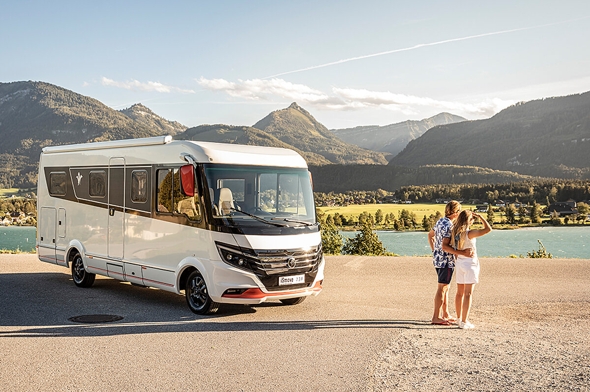 Ein paar steht neben einem großen Wohnmobil und blickt auf einen See.