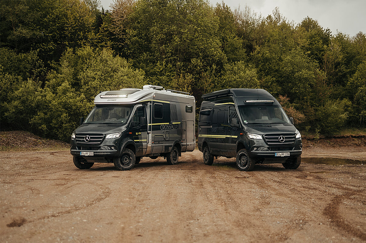 Zwei Abenteuer-Camper-Vans stehen auf einer sandigen Straße vor einem Wald.