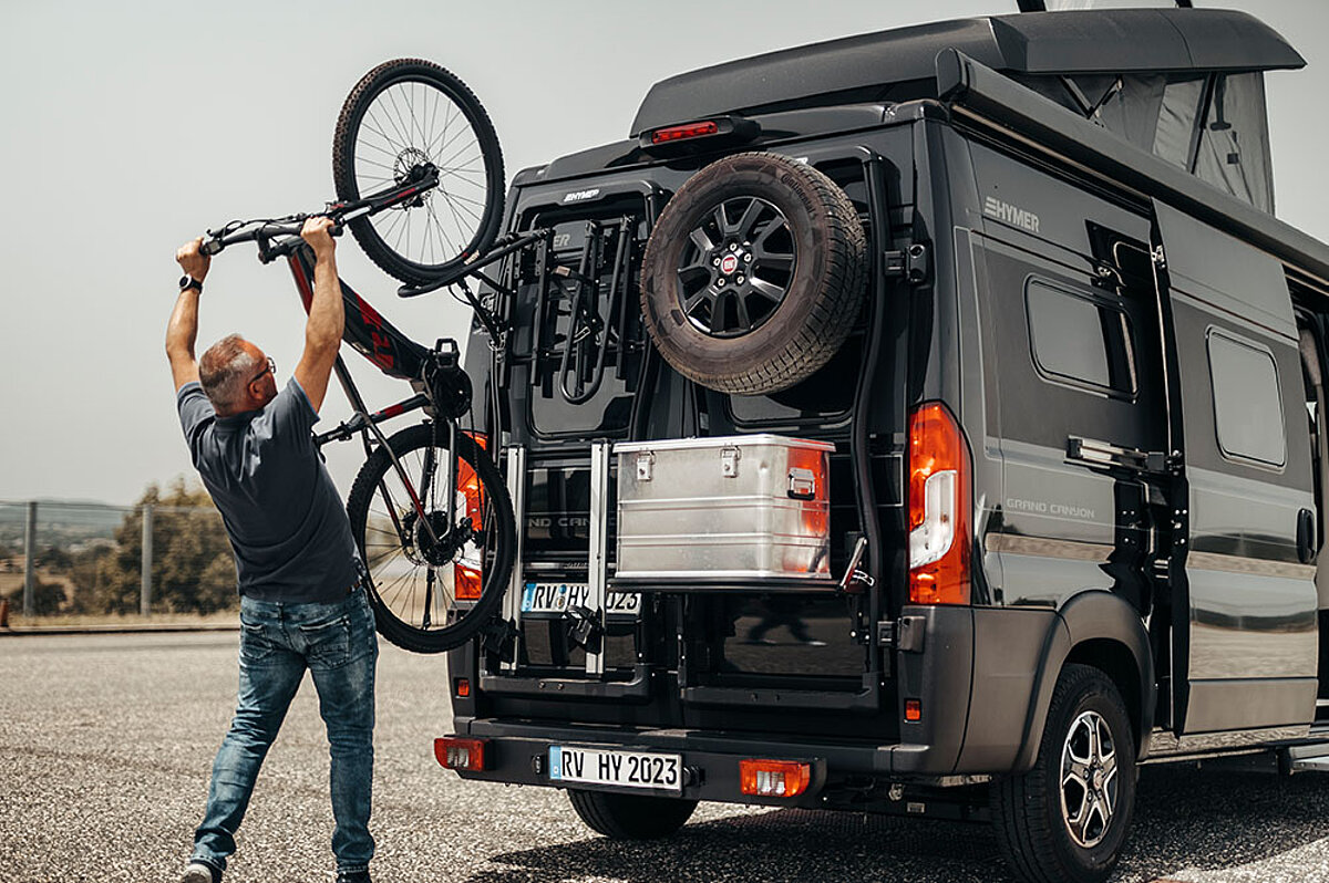 Wir rüsten Ihr Fahrzeug für jedes Abenteuer nach. Ein Mann nimmt ein Fahrrad vom Heckträger eines Camper Vans.