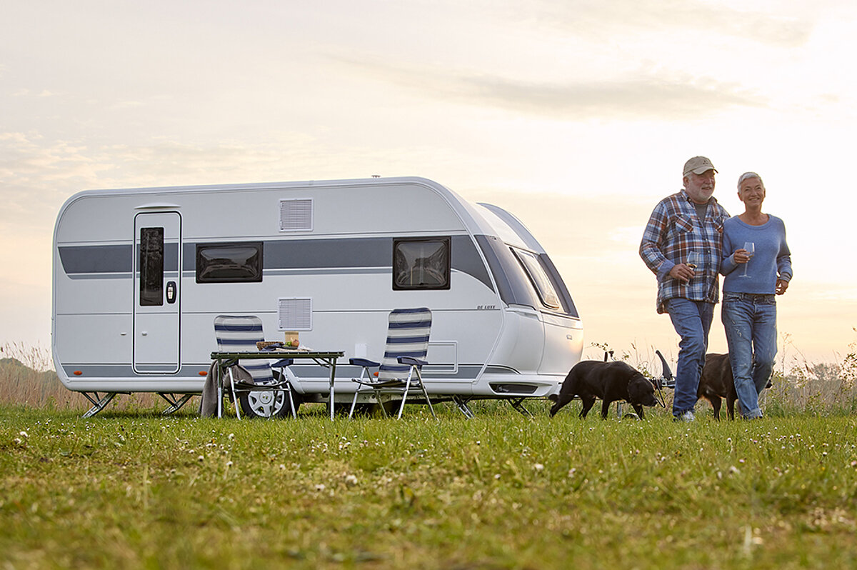 Unbeschwert Urlaub machen – dank Ihrer Wohnwagen-Versicherung! Ein älteres Paar läuft mit zwei Hunden über die Wiese vor einem Wohnwagen.