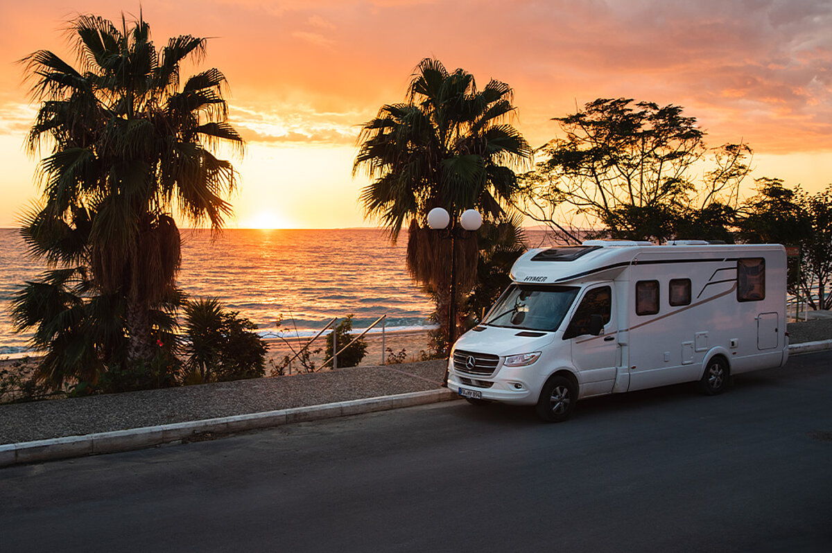 Ein Wohnmobil fährt eine Straße neben Palmen, Strand und Sonnenuntergang entlang.