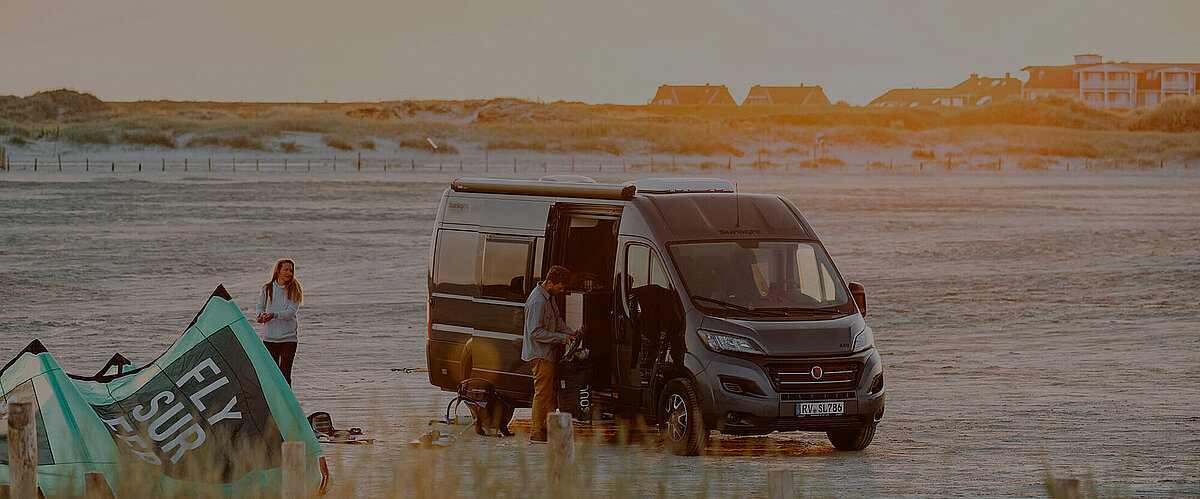 Zwei junge Menschen bauen am Strand ein Zelt neben ihrem Camper Van auf.