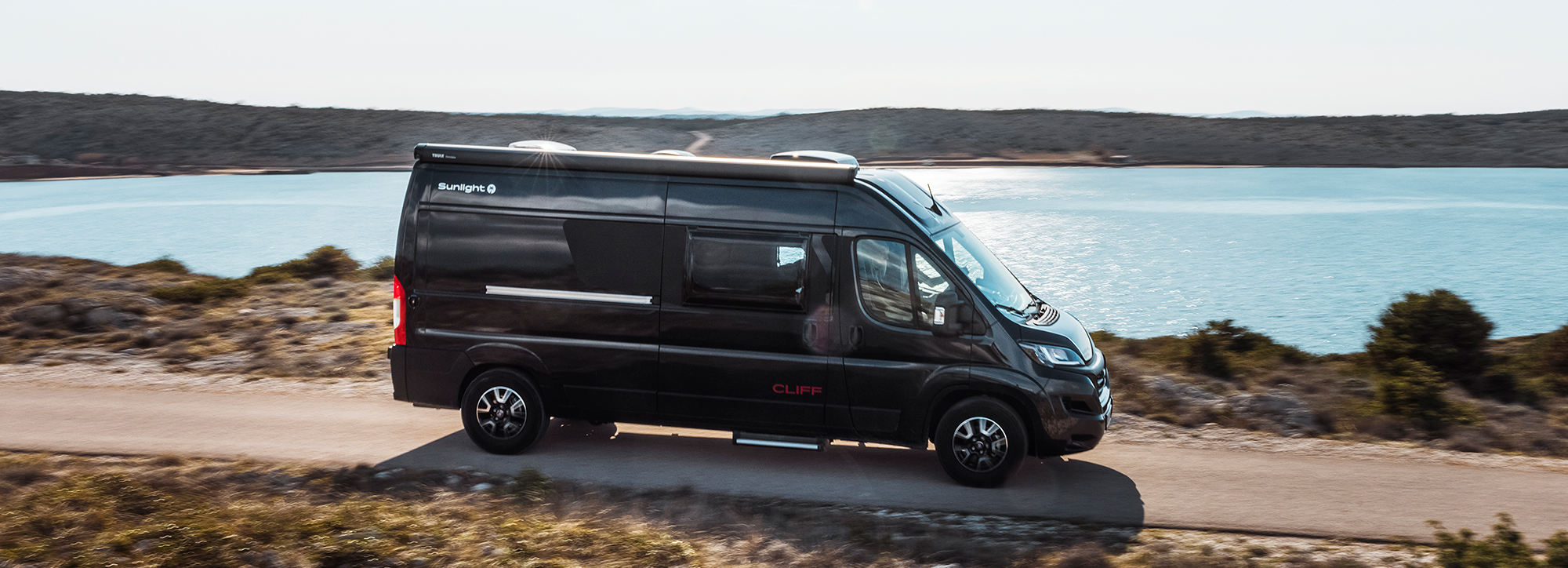 Ein schwarzer Campervan fährt über eine Küstenstraße.