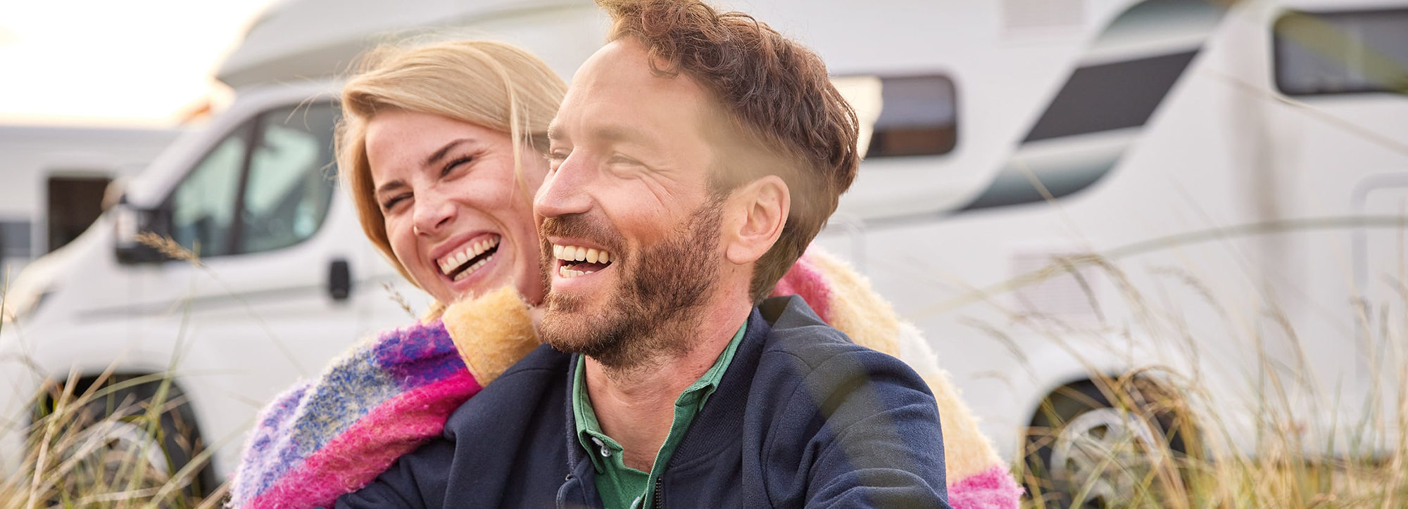 Ein Paar sitzt im Gras und lacht, hinter ihnen stehen zwei Wohnmobile.