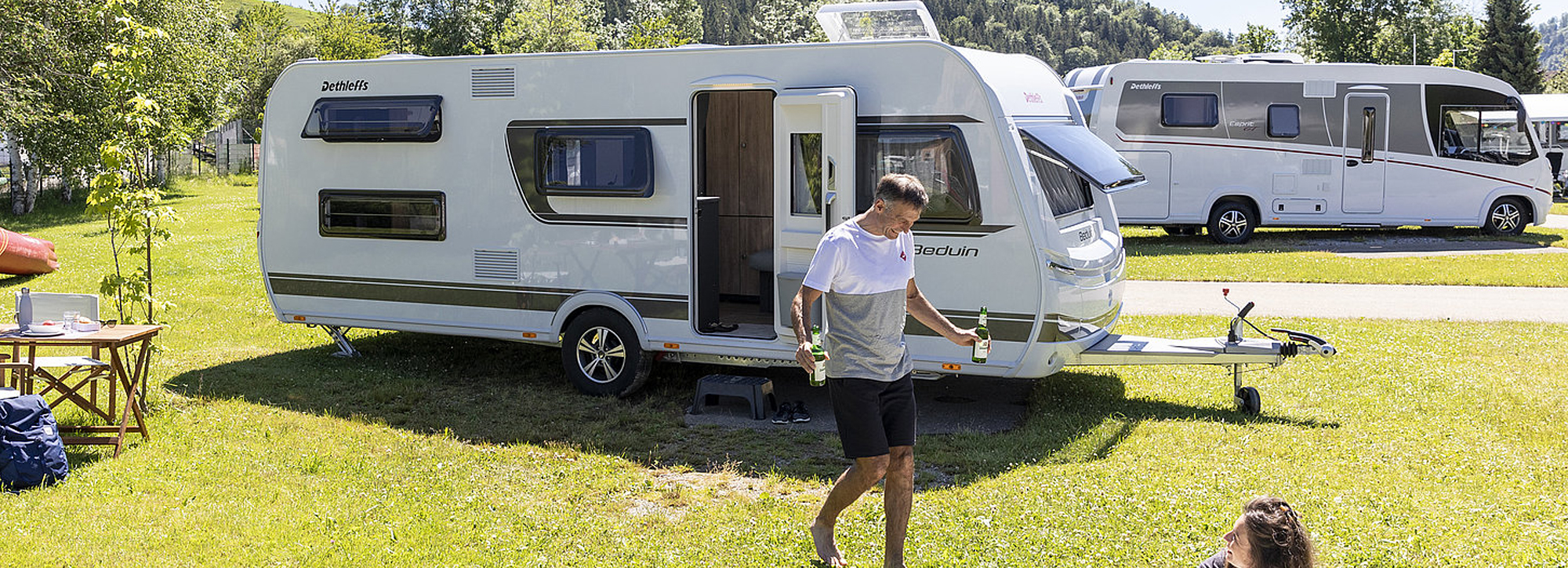 Ein Mann bringt seiner Frau, die vor dem Caravan auf der Wiese sitzt, ein Bier.